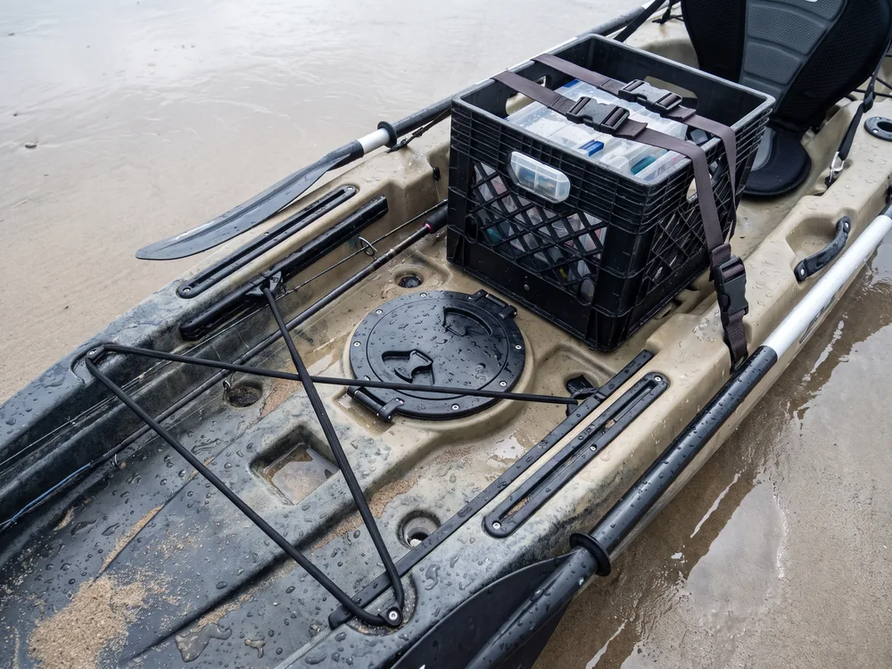 Overhead view of a rigged kayak deck on wet sand with the crate strapped down, the hatch closed and latched, rods lying secured in horizontal holders and the paddle leashed along the gunwale