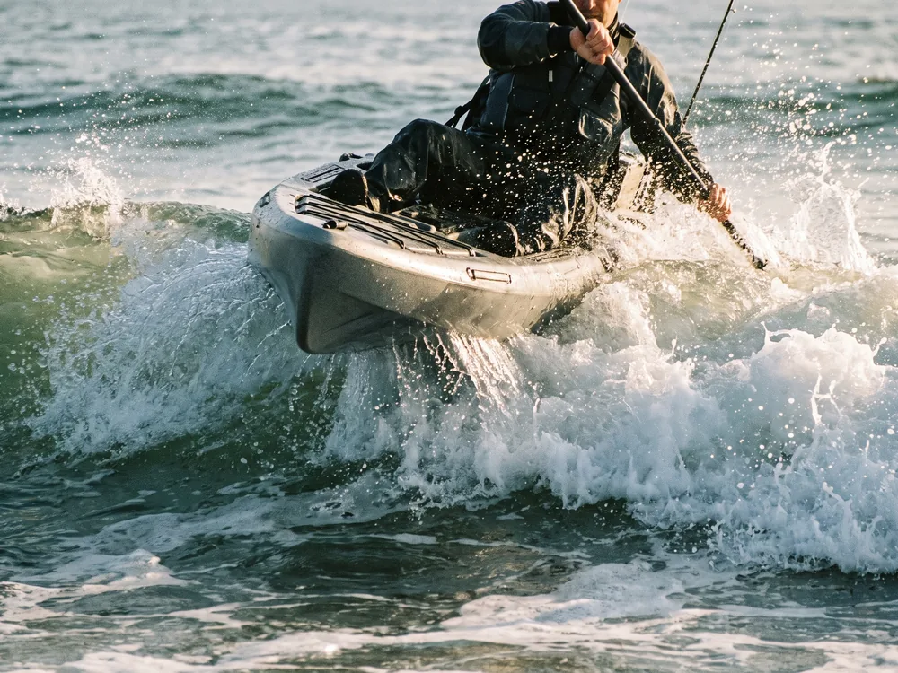 A sit-on-top fishing kayak driving bow-first through a breaking wave, spray coming over the front deck