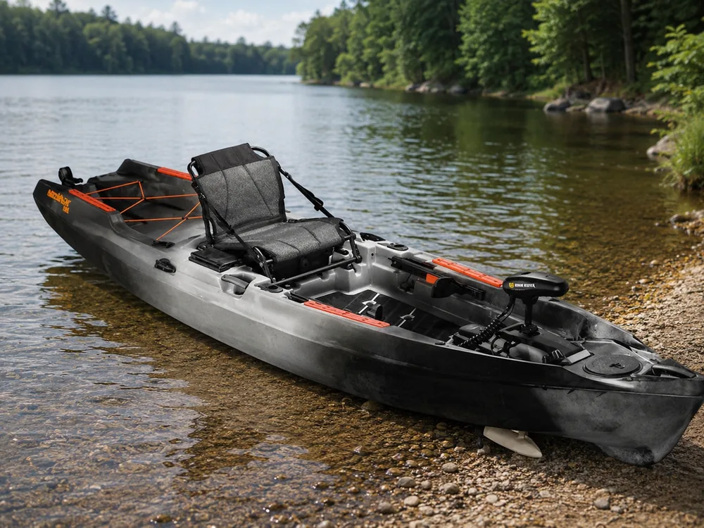 A black and grey fishing kayak on a lake shore with a stern-mounted trolling motor and its coiled power lead running into the hull