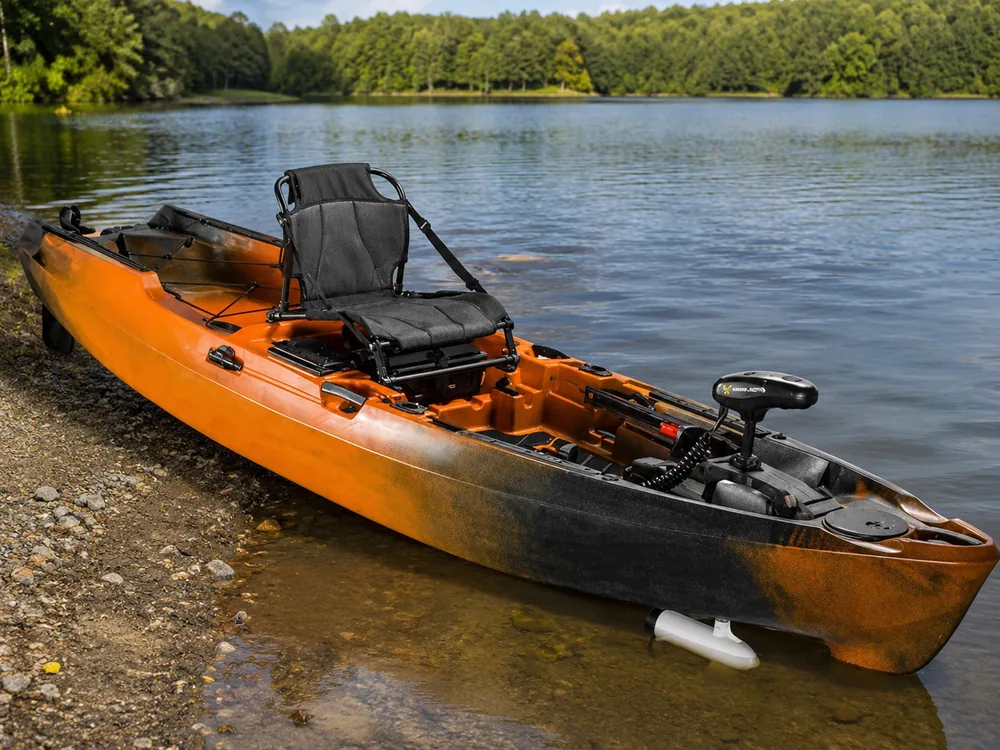 An orange and black fishing kayak resting on a gravel shore with a stern-mounted trolling motor fitted