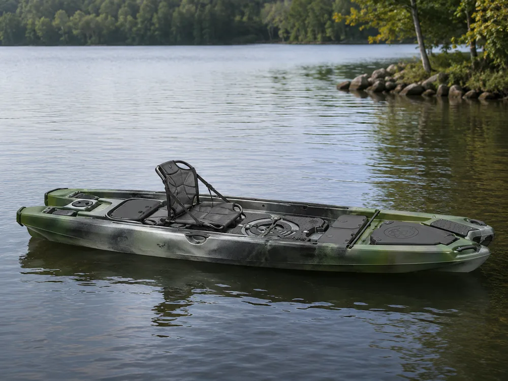 A camouflage fishing kayak with a flat open deck sitting on calm open water beside a wooded shoreline