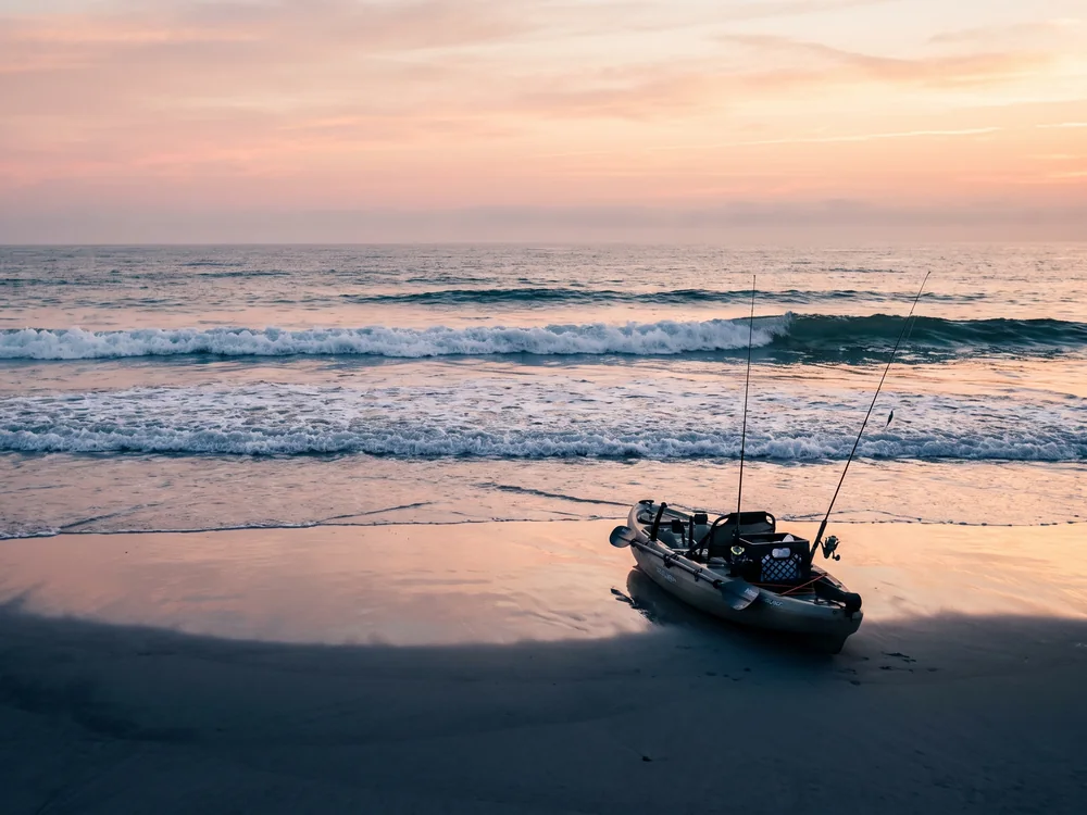 A rigged fishing kayak resting at the waterline at dawn with clean lines of small surf breaking behind it
