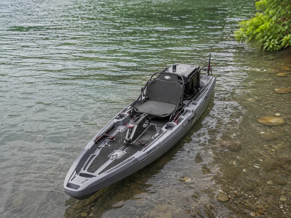 A rigged fishing kayak floating in clear shallow water beside a green shoreline